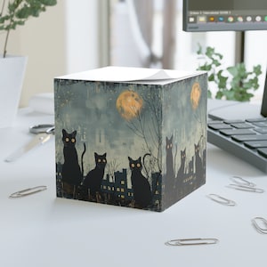 May include: A cube-shaped notepad dispenser featuring a spooky Halloween design. The artwork depicts black cats with glowing yellow eyes silhouetted against a night scene with a full moon, trees, and buildings. The top of the cube holds a stack of white notepaper.