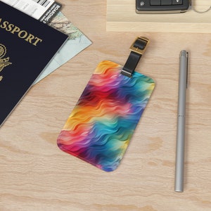 May include: A colorful luggage tag with a wavy, abstract pattern in shades of red, orange, yellow, green, blue, and purple. The tag has a black leather strap with a gold buckle.