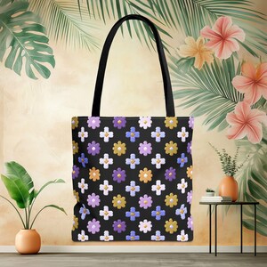 May include: Black tote bag with a repeating floral pattern in white, purple, yellow, and blue. The bag has black handles and a black background. The bag is made of a durable material and is suitable for carrying groceries or other items.