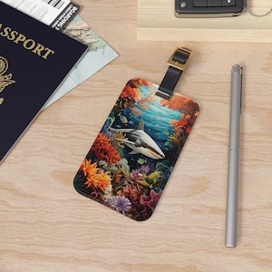 May include: A colorful luggage tag with a white shark swimming through a coral reef. The tag has a black leather strap with a gold buckle.