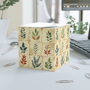May include: A square notepad with a beige background and a collage of different green, blue, and red leaves. The notepad has a white top sheet.