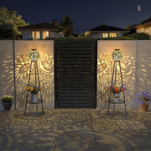 May include: Two black metal solar powered lanterns with intricate cut-out designs of suns and moons. The lanterns are on tall stands and cast shadows on a white wall. The lanterns are lit and glowing.