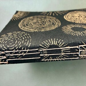 May include: A handmade journal with a black cover featuring a repeating pattern of beige concentric circles and radial lines. The binding is visible, with white stitching along the spine. The journal is square and appears to be new.