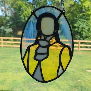 May include: Stained glass oval depicting a figure in a yellow coat and white cravat, suspended by a silver chain. The figure's face is blank. The glass is framed in black, with a background of blue and green. The piece is hanging outdoors.