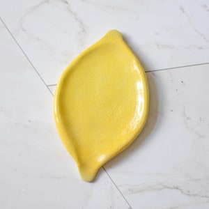 K&ouml;nnte beinhalten: Eine leuchtend gelbe Keramikschale in Zitronenform. Die Oberfl&auml;che hat ein gl&auml;nzendes, leicht strukturiertes Finish mit einem spitzen Ende an der Oberseite und einer kleinen Vertiefung an der Unterseite, die das Aussehen einer Zitrusfrucht nachahmt.