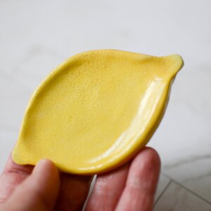 K&ouml;nnte beinhalten: Eine kleine, leuchtend gelbe Keramikschale in Form einer Zitrone. Die Oberfl&auml;che der Schale weist ein texturiertes, genopptes Finish auf, das das Aussehen einer echten Zitronenschale nachahmt, mit glatten, leicht erh&ouml;hten R&auml;ndern und einem spitzen Ende.
