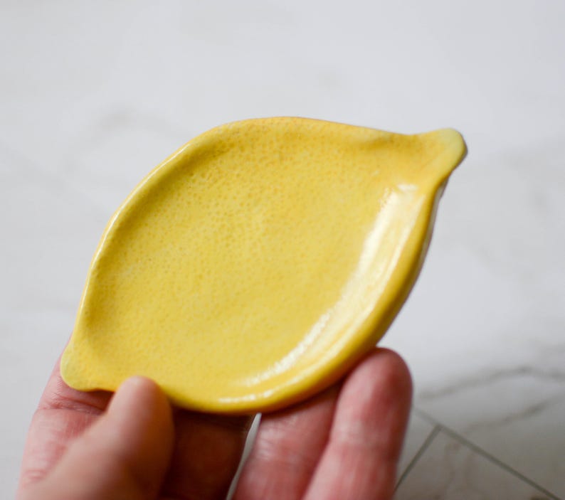 K&ouml;nnte beinhalten: Eine kleine, leuchtend gelbe Keramikschale in Form einer Zitrone. Die Oberfl&auml;che der Schale weist ein texturiertes, genopptes Finish auf, das das Aussehen einer echten Zitronenschale nachahmt, mit glatten, leicht erh&ouml;hten R&auml;ndern und einem spitzen Ende.