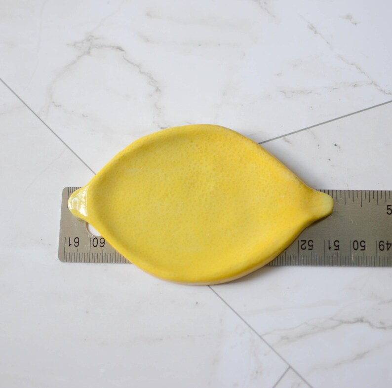 K&ouml;nnte beinhalten: Eine gelbe, zitronenf&ouml;rmige Keramikschale, die auf einem Metalllineal liegt. Das Lineal zeigt, dass die Schale etwa 11,5 cm lang ist und sich von der 52- bis zur 60,5-Markierung auf der Skala erstreckt, mit einer strukturierten Oberfl&auml;che.