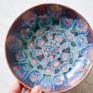 May include: A round ceramic bowl featuring a vibrant crystalline glaze with a repeating pattern of blue, purple, and golden-yellow petal-like shapes radiating from the center. The rim and exterior base of the bowl show a warm, natural terracotta clay color.
