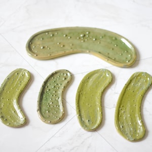 Könnte beinhalten: Ein Set aus fünf Keramikschalen in Form von Gewürzgurkenscheiben mit einer glänzenden hellgrünen Glasur und erhabenen Noppen auf der Oberfläche. Eine große, gebogene Schale liegt oben, darunter vier kleinere, längliche, ovale Schalen.