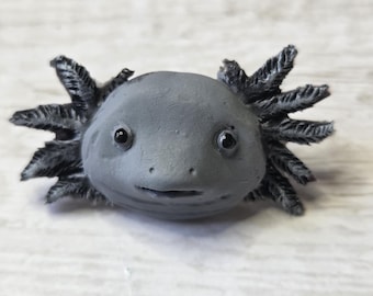 May include: A gray axolotl figurine with black feathery gills and small, dark eyes. The axolotl has a smooth, rounded head and a slight smile. The figurine is a detailed representation of the aquatic salamander.