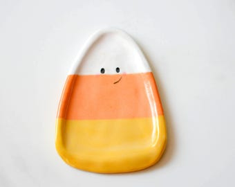 Könnte beinhalten: Eine Keramikschale in Form eines Zuckermais-Bonbons. Die Schale hat drei horizontale Farbbänder: Weiß oben, Orange in der Mitte und Gelb unten. Ein einfaches Gesicht mit zwei schwarzen Augen und einem kleinen Lächeln ist auf dem weißen Bereich aufgemalt.