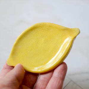 K&ouml;nnte beinhalten: Eine leuchtend gelbe Keramikschale in Zitronenform. Die Oberfl&auml;che der Schale weist eine texturierte, genoppte Struktur auf, die der Schale einer Zitrone nachempfunden ist, mit einer glatten, gl&auml;nzenden Oberfl&auml;che und spitz zulaufenden Enden.