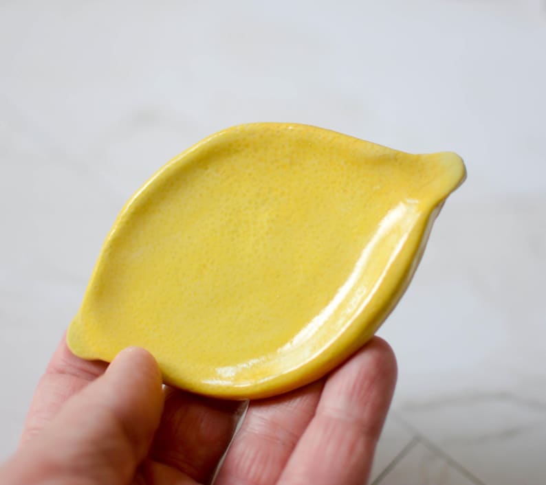 K&ouml;nnte beinhalten: Eine leuchtend gelbe Keramikschale in Zitronenform. Die Oberfl&auml;che der Schale weist eine texturierte, genoppte Struktur auf, die der Schale einer Zitrone nachempfunden ist, mit einer glatten, gl&auml;nzenden Oberfl&auml;che und spitz zulaufenden Enden.