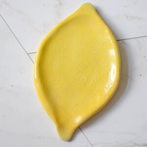 K&ouml;nnte beinhalten: Eine leuchtend gelbe Keramik-L&ouml;ffelablage in Zitronenform. Die Oberfl&auml;che hat ein gl&auml;nzendes, strukturiertes Finish mit einer leicht spitz zulaufenden Oberseite und einer kleinen, abgerundeten Vertiefung an der Unterseite, die die Form einer Zitrone nachahmt.