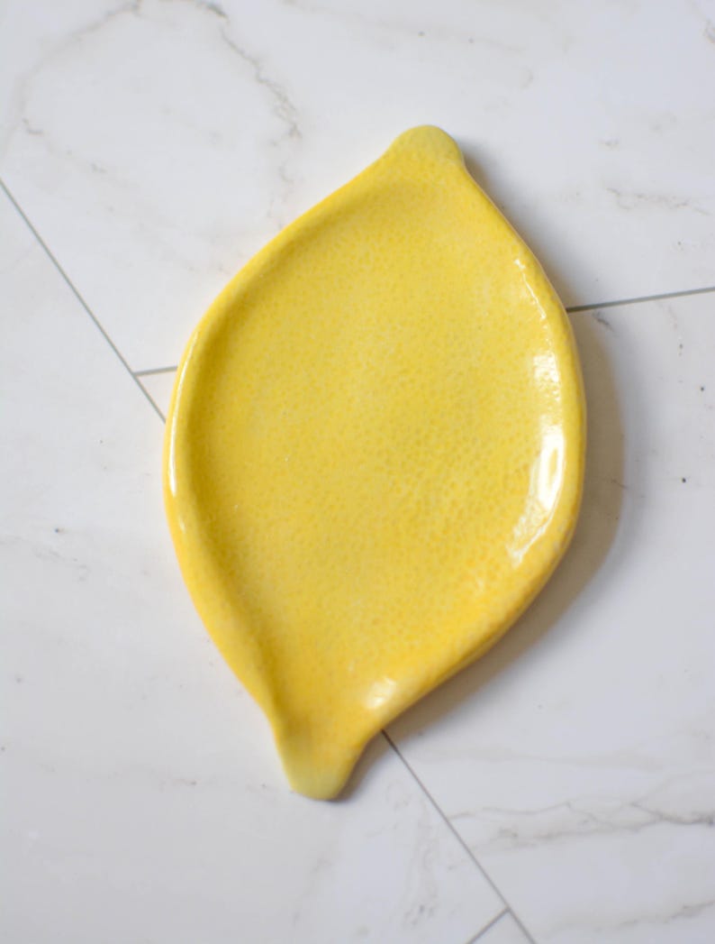 K&ouml;nnte beinhalten: Eine leuchtend gelbe Keramik-L&ouml;ffelablage in Zitronenform. Die Oberfl&auml;che hat ein gl&auml;nzendes, strukturiertes Finish mit einer leicht spitz zulaufenden Oberseite und einer kleinen, abgerundeten Vertiefung an der Unterseite, die die Form einer Zitrone nachahmt.
