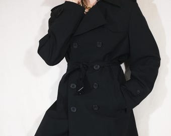 Op de afbeelding: Een zwarte trenchcoat met een dubbele rij knopen, met een riem in de taille en manchetten met knopen. De jas heeft een klassieke kraag en zakken. Het model draagt een blauwe jeans.