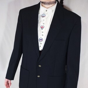 May include: A navy blue blazer with two front pockets and gold buttons. The blazer is worn over a white blouse with embroidered patches and a red and black plaid skirt.