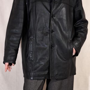 May include: A black leather jacket with a notched collar and three-button closure. The jacket has two front pockets and is worn over a gray turtleneck sweater and gray pants. The jacket is a classic style.