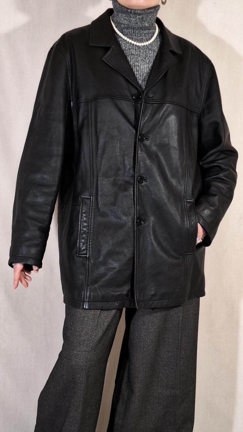 May include: A black leather jacket with a notched collar and three-button closure. The jacket has two front pockets and is worn over a gray turtleneck sweater and gray pants. The jacket is a classic style.