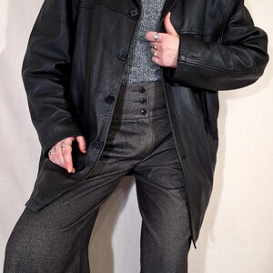 May include: A black leather coat with a button closure, worn over a gray turtleneck sweater and high-waisted gray trousers. The trousers have a button fly. A pearl necklace and rings are also visible.