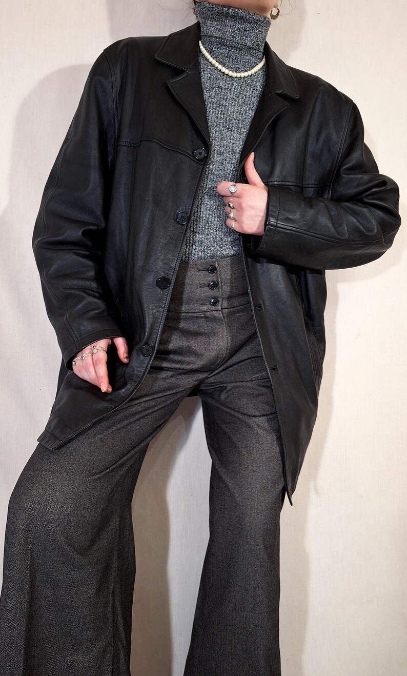 May include: A black leather coat with a button closure, worn over a gray turtleneck sweater and high-waisted gray trousers. The trousers have a button fly. A pearl necklace and rings are also visible.
