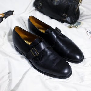 May include: Two black leather monk strap shoes with gold buckles on a white surface.