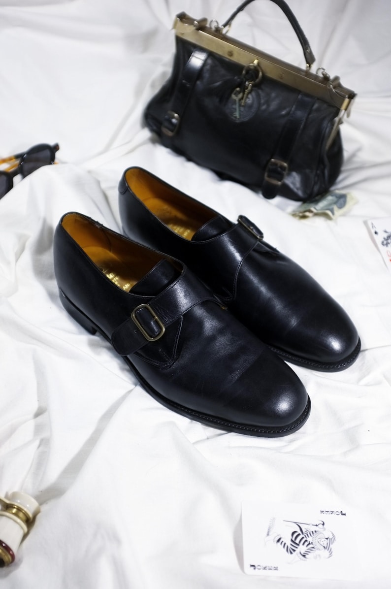 May include: Two black leather monk strap shoes with gold buckles on a white surface.