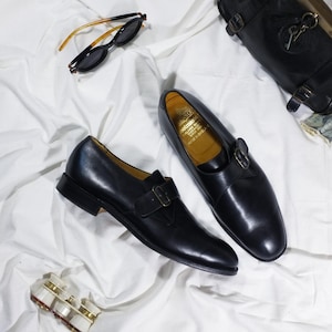 May include: A pair of black leather monk strap shoes with a gold buckle. The shoes are on a white fabric background.