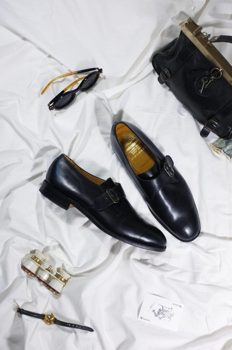 May include: A pair of black leather monk strap shoes with a gold buckle. The shoes are on a white fabric background.