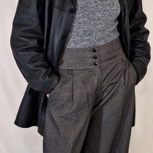 May include: A black leather jacket over a gray turtleneck sweater and high-waisted gray trousers. The trousers have a buttoned closure and pleats. A pearl necklace is visible.