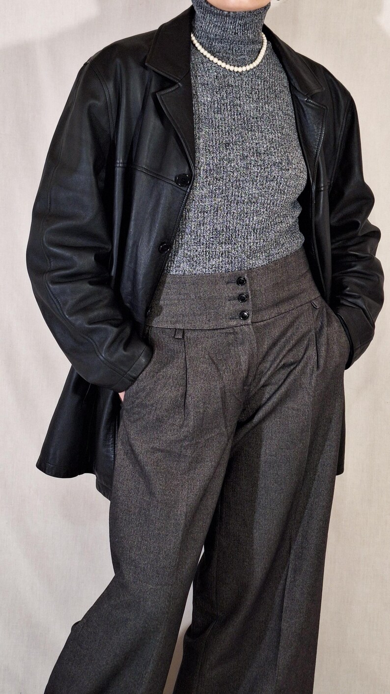 May include: A black leather jacket over a gray turtleneck sweater and high-waisted gray trousers. The trousers have a buttoned closure and pleats. A pearl necklace is visible.