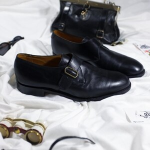 May include: Two black leather monk strap shoes with gold buckles on a white fabric background.