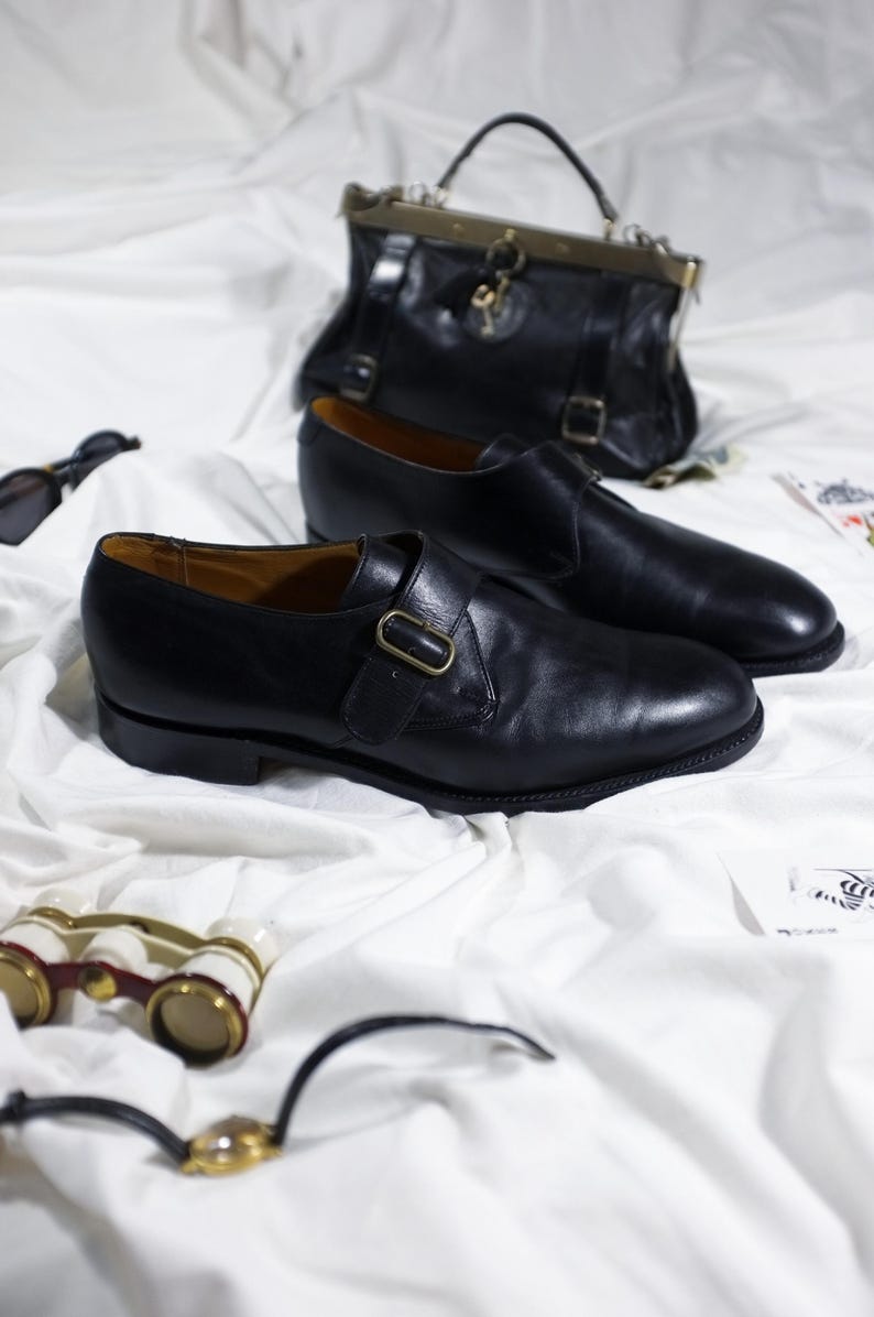 May include: Two black leather monk strap shoes with gold buckles on a white fabric background.