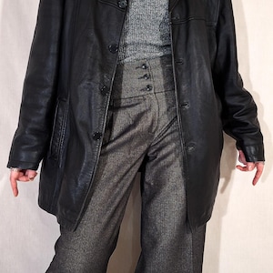May include: A black leather coat with a button-up front, worn over a gray turtleneck sweater and high-waisted gray trousers. The outfit is completed with a pearl necklace.