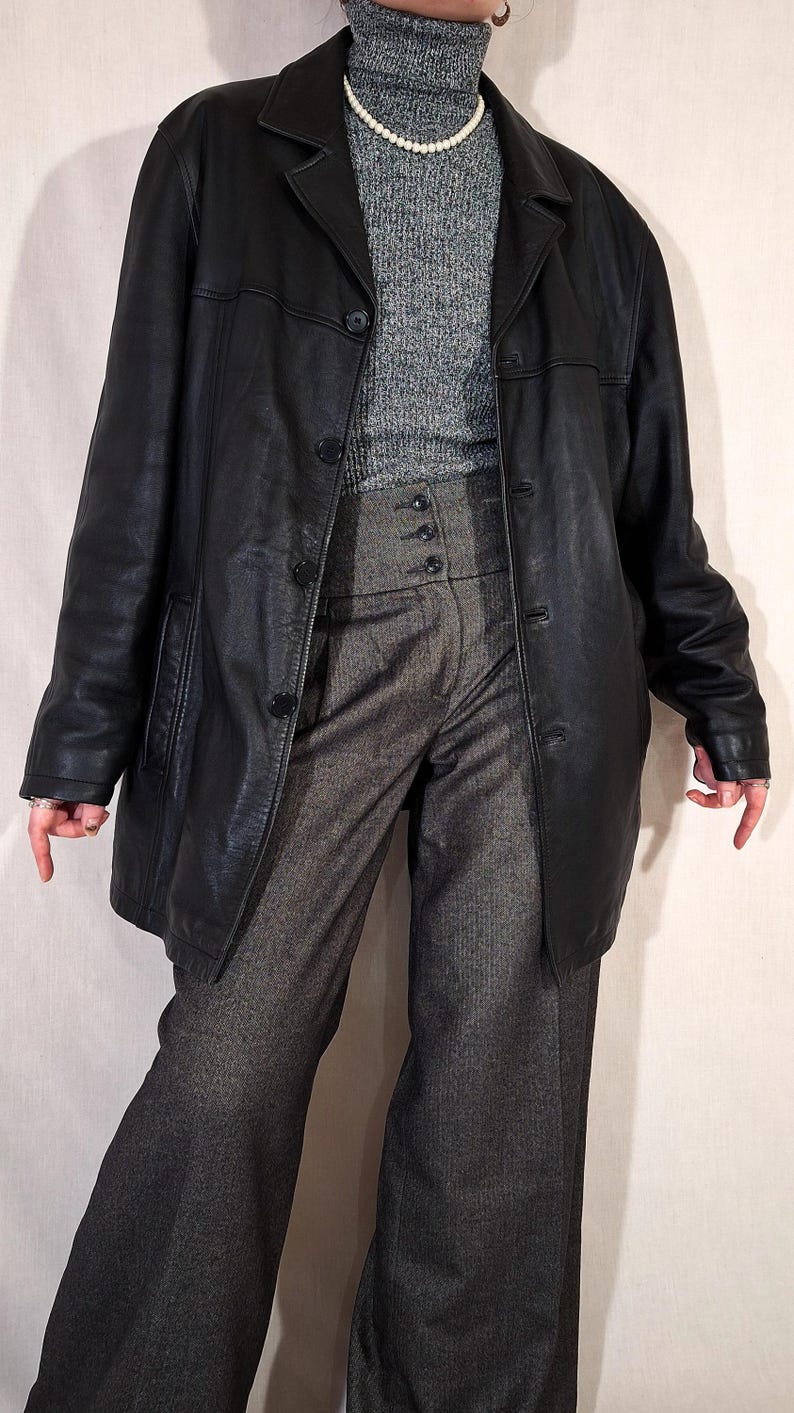 May include: A black leather coat with a button-up front, worn over a gray turtleneck sweater and high-waisted gray trousers. The outfit is completed with a pearl necklace.