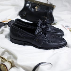 May include: A pair of black leather loafers with a tassel detail and laces. The shoes are on a white surface.