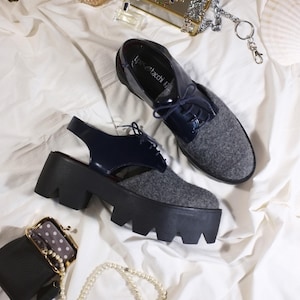 Pode incluir: Sapatos de plataforma femininos com cabedais de feltro cinza e detalhes em couro envernizado azul marinho. Os sapatos têm solas pretas grossas e são exibidos com uma pequena bolsa preta, joias e frascos de perfume em uma superfície branca.