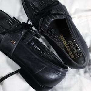 May include: A pair of black leather tassel loafers with black laces. The loafers have a  perforated design on the inside and the text "Regal Obermaier" is printed on the insole.