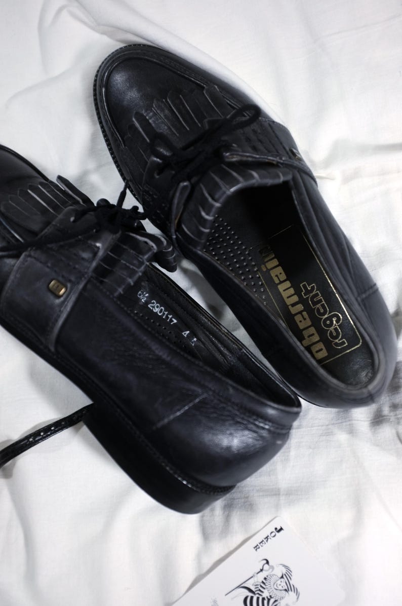 May include: A pair of black leather tassel loafers with black laces. The loafers have a  perforated design on the inside and the text "Regal Obermaier" is printed on the insole.
