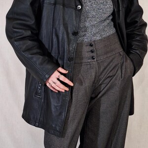 May include: A black leather blazer with a classic collar and button closure. The blazer is paired with a gray turtleneck sweater, high-waisted gray trousers, and a pearl necklace. The trousers have a buttoned waist detail.