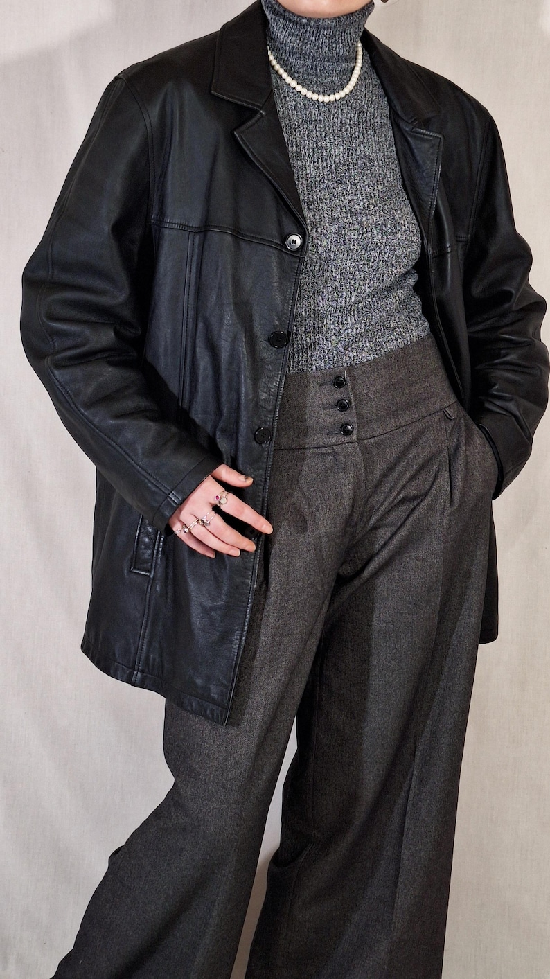May include: A black leather blazer with a classic collar and button closure. The blazer is paired with a gray turtleneck sweater, high-waisted gray trousers, and a pearl necklace. The trousers have a buttoned waist detail.