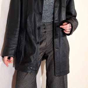 May include: A black leather jacket with a button-up front, worn over a gray turtleneck sweater and high-waisted gray trousers. The jacket has a classic lapel and pockets. The outfit is accessorized with a pearl necklace and rings.