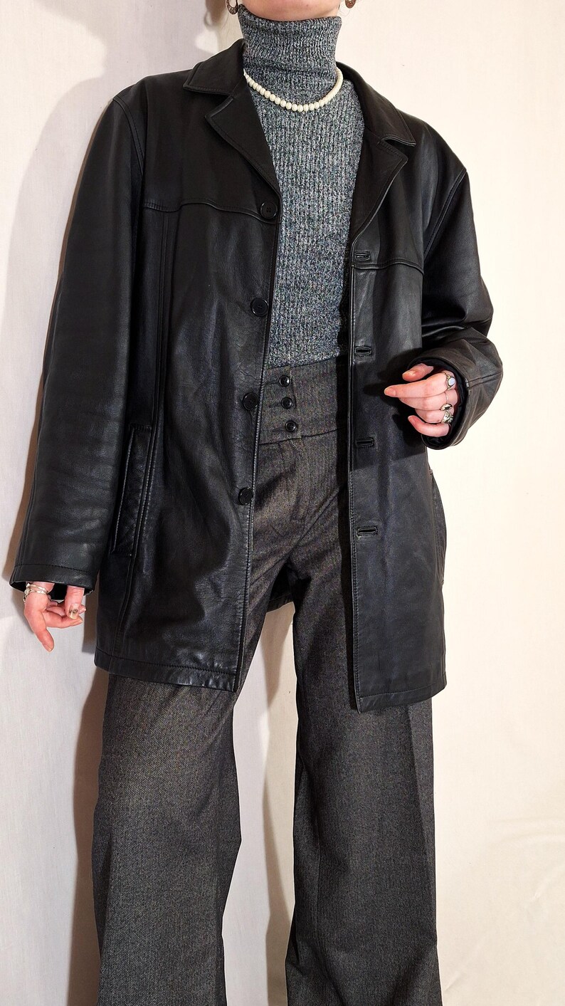 May include: A black leather jacket with a button-up front, worn over a gray turtleneck sweater and high-waisted gray trousers. The jacket has a classic lapel and pockets. The outfit is accessorized with a pearl necklace and rings.