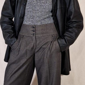 May include: A black leather jacket over a gray turtleneck sweater and gray high-waisted trousers. The trousers have a buttoned waistband and pleats. A pearl necklace is visible.