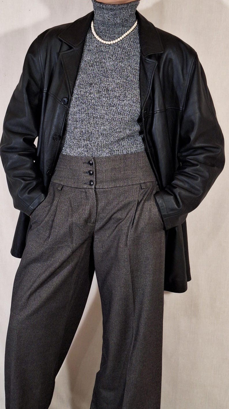 May include: A black leather jacket over a gray turtleneck sweater and gray high-waisted trousers. The trousers have a buttoned waistband and pleats. A pearl necklace is visible.