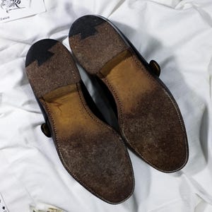 May include: A pair of black leather shoes with a brown leather sole. The shoes have a buckle strap and a pointed toe.