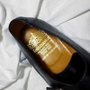 May include: Close-up of the inside of a black leather shoe with a gold embossed label that reads "Grenson footmaster Englands Master Shoemaker Since 1866 Made in England."
