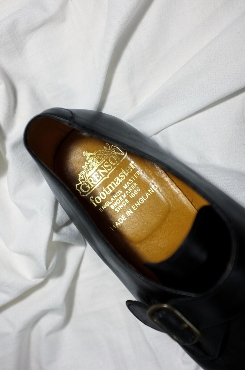May include: Close-up of the inside of a black leather shoe with a gold embossed label that reads "Grenson footmaster Englands Master Shoemaker Since 1866 Made in England."
