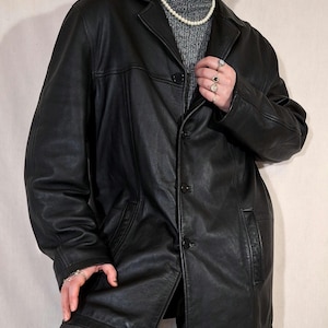 May include: A black leather jacket with a classic design. The jacket features a collared neckline, button-down closure, and front pockets. The jacket is worn over a gray turtleneck sweater and gray pants.